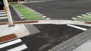 Closeup view of an intersection corner on the Scott Street cycle track. The track continues from left to right, and a crossride painted green connects from across the street. Tactile pavers delineate the sidewalk from the asphalt cycle track.