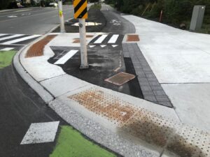 The beginning of a cycle track at an intersection. The track runs over a shaved concrete curb with embedded tactile plates and then continues as an asphalt path next to the sidewalk. There is a utility pole on the left side within the width of the track, and painted markings indicate cyclists turning left should wait obstructing the track.