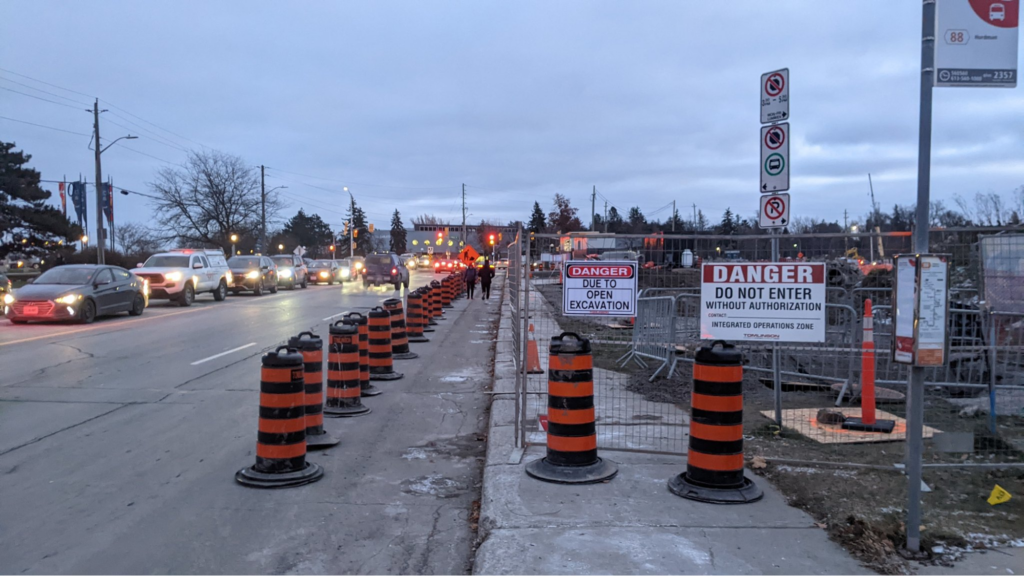 A sidewalk blocked by construction fencing, and a line of traffic cones has been placed about 2 metres from the curb blocking the lane and separating a walkway from traffic.