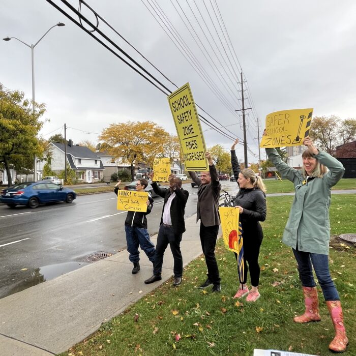 Parents and councillors rally to keep speed safety cameras in front of Queen Elizabeth Public School. They’re holding signs that read: school safety zone, safer school zones, honk to support speed safety cameras