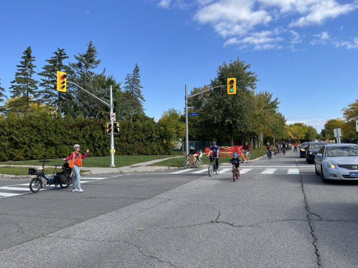 Lots of acults and kids riding on Jeanne D’Arc Boulevard as part of a kidical mass ride to demonstrate the need for safe cycling infrastructure. They’re taking a lane of traffic. To their right, people in cars.