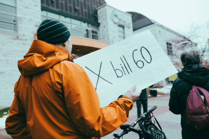 Protesters seen from the back at Bill 60 rally. In the foreground, one is holding a sign that reads: X bill 60