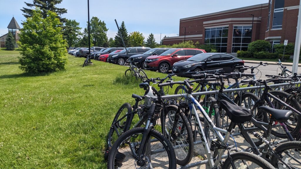 Many bikes parked and leaning at awkward angles at a schoolyard rack. Bikes on signposts with only one way to lock are in the background.