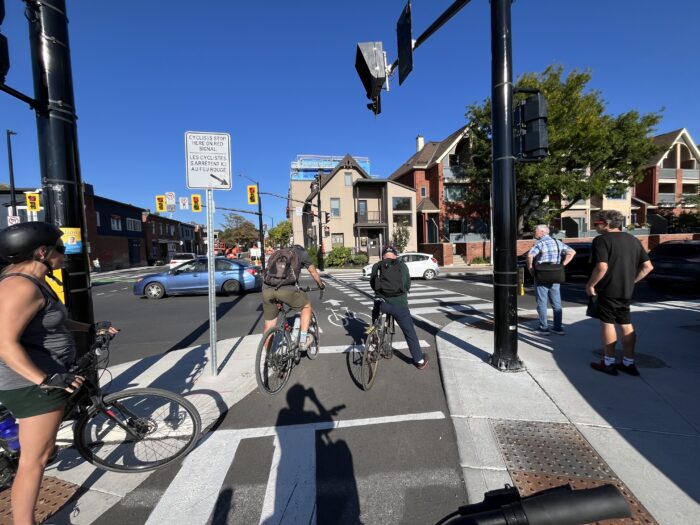 Cyclists and pedestrians waiting at their respective crossride and crosswalk at the red light. Towards the top left corner, one can see a sign banning right-on-red turns.